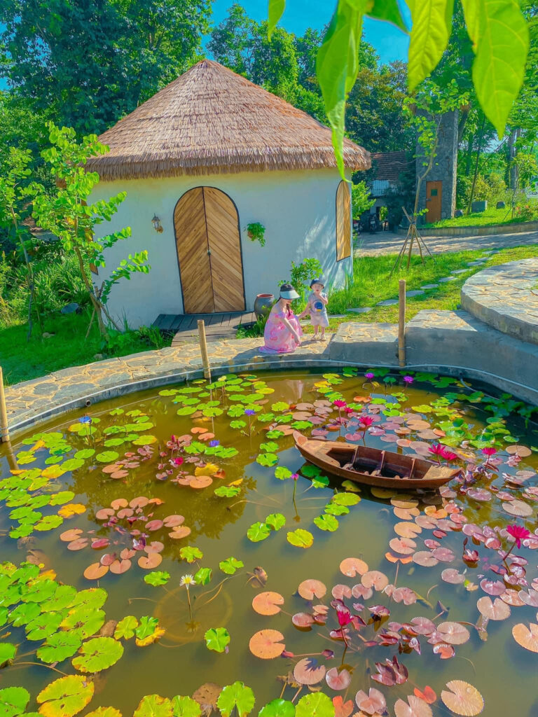 Ngôi nhà mái lá sử dụng rơm nhân tạo làm mái, nằm cạnh hồ sen và bao quanh bởi cây xanh.