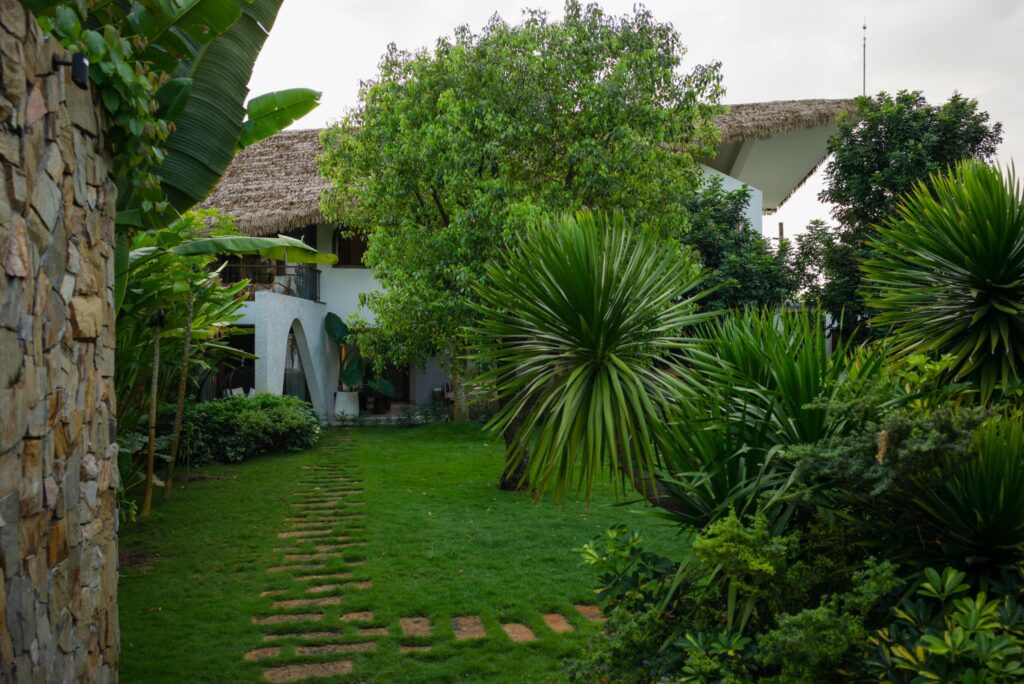 Lối đi lát đá tự nhiên dẫn vào căn nhà sử dụng mái rơm nhân tạo, tạo cảm giác thân thiện và gần gũi thiên nhiên.