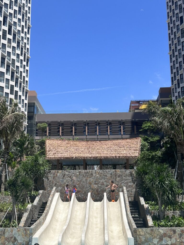 Cầu trượt hồ bơi resort nằm giữa hai tòa nhà cao tầng, phía trên là chòi nghỉ lợp mái lá nhân tạo.