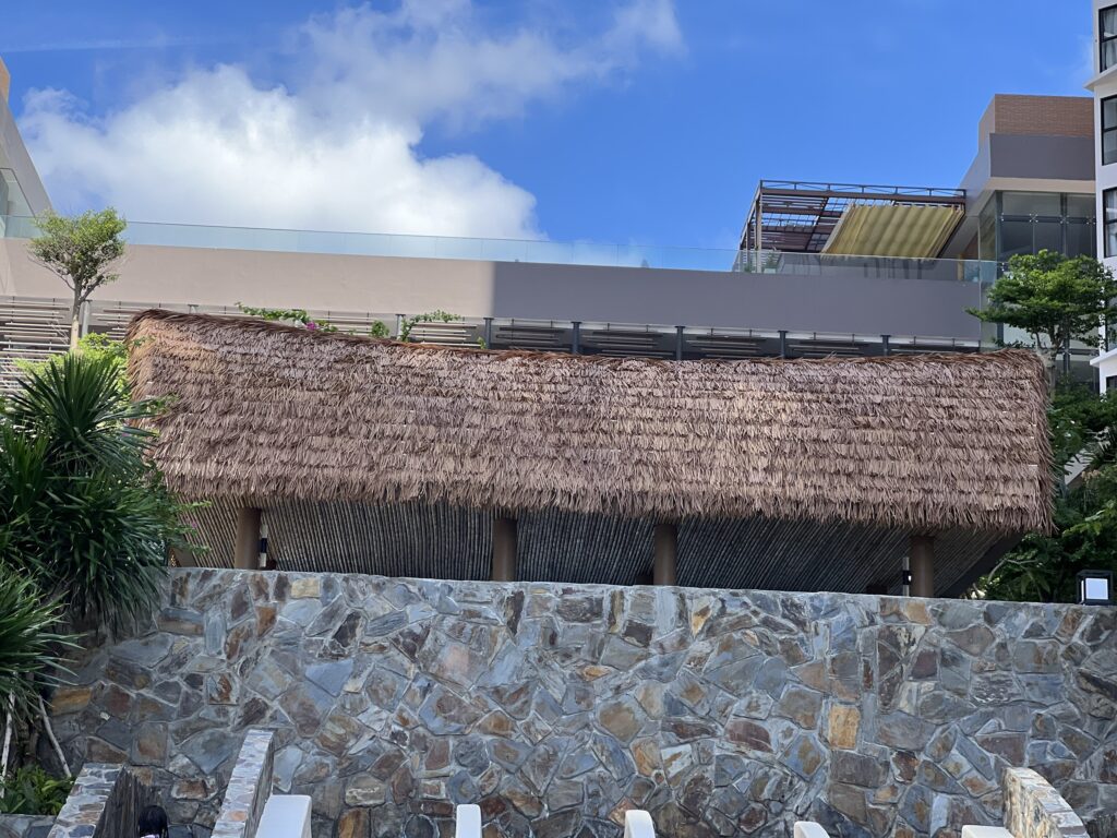Chòi nghỉ lớn lợp mái lá nhân tạo tại khu nghỉ dưỡng, phía trước là bức tường đá và hàng ghế hồ bơi.