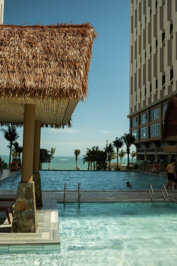 Mái chòi hồ bơi resort lợp mái lá nhân tạo hướng biển với khung cột chắc chắn, bên cạnh tòa nhà cao tầng.